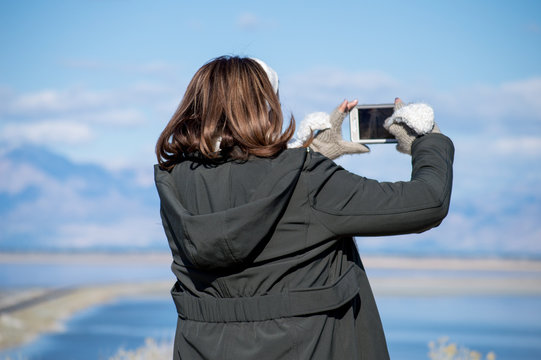 young woman taking picture of scenery view of blue lake and mountains picture with cell phone wearing winter jacket and gloves