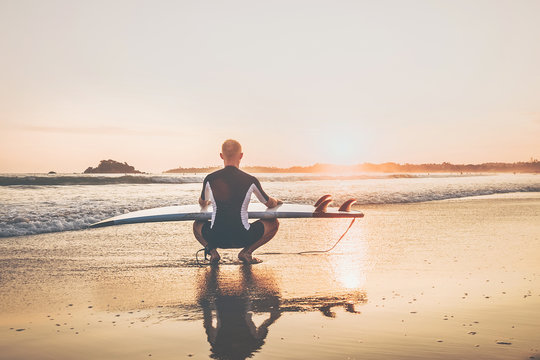 Surfer man with long board surf sitting on the sandy ocean beach and enjoying the sunset sky. Never ending summer surfing concept image.