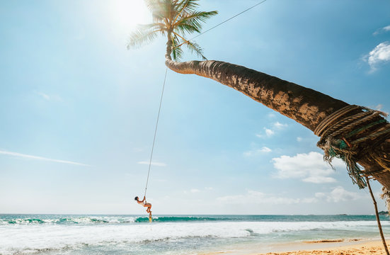 Woman in white swimsuit swinging on tropical palm swing over the ocean waves. Careful summer tropic climate countries vacation concept image.