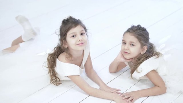 Two beautiful little girls posing for a photographer. They lie on the floor, chatter legs and look into the camera. On children for white white fluffy dresses, like those of the bride