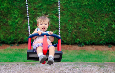Cute scared little boy riding on swing with panicked  face expression at children playground on summer day. Child emotions. Copy space.