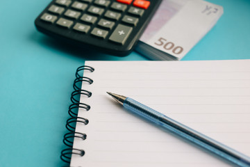 clear notebook, pen, pack of Euro banknotes and a calculator on a blue background