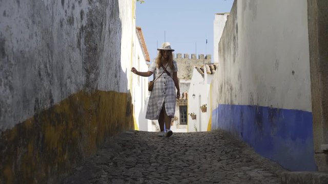 Pretty young woman walking on narraow street between building. Girl going on camera smiling , touching wall and enjoying sunny day. Obidos, Portugal