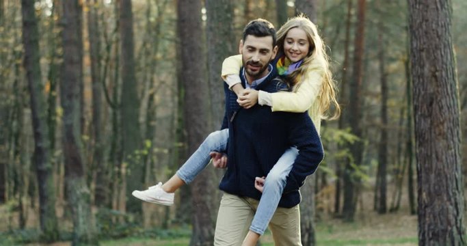 Attractive Caucasian father walking the forest and carrying his cute small daughter on his back while they talking. Outdoors.