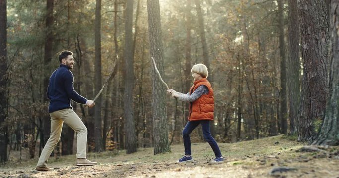 Cheerful Caucasian father and little son playing in the forest with sticks like they fighting with swords. Outdoor.