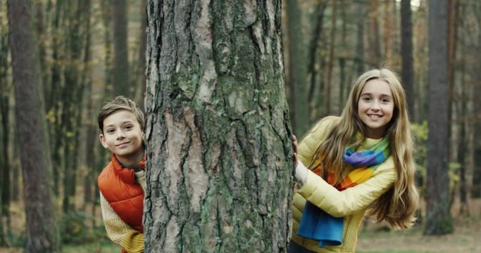 Portrait shot of the cheerful cute boy and girl standing behind the tree and peering from its trunk while smiling to the camera. Outside.