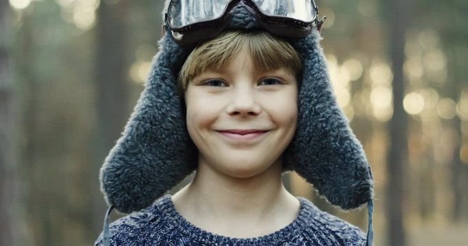 Close up of the small cute Caucasian boy dreamer of being a pilot in the hat with glasses smiling cheerfully to the camera in the forest or a park. Portrait. Outdoor.