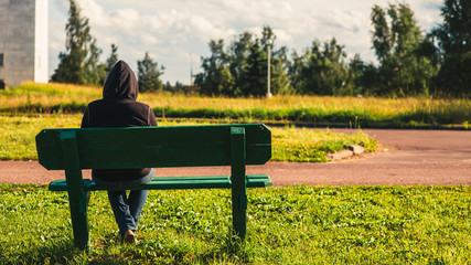The lonely man sits on a bench in a hoodie overlooking the green park landcape full of trees and...