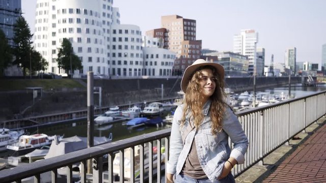 Pretty girl tourist in casual wear standing on embankment at background yacht in harbor and modern buildings. Crop woman with photo camera and holding smartphone in hand. Dusseldorf, Germany