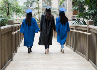 Three girl friends graduate walking on bridge wearing blue black graduation cap and gown holding...