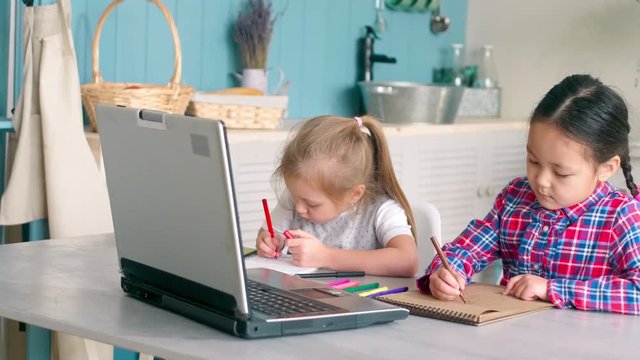 Cute Caucasian and Asian little girls sitting together at kitchen table at home, looking at laptop screen and drawing pictures in sketchbooks with felt tip pens