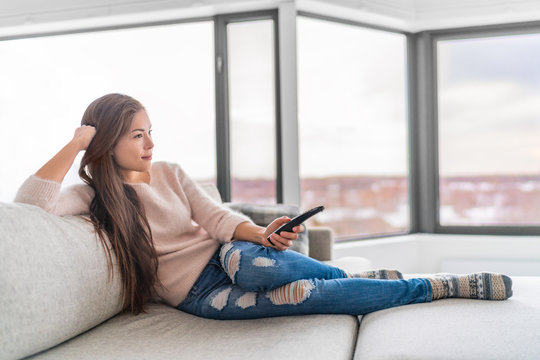 Woman Watching TV At Home Sitting On Sofa In Modern Apartment. Young Casual Asian Girl Enjoying TV Show Holding Remote. Comfort Living Lifestyle.