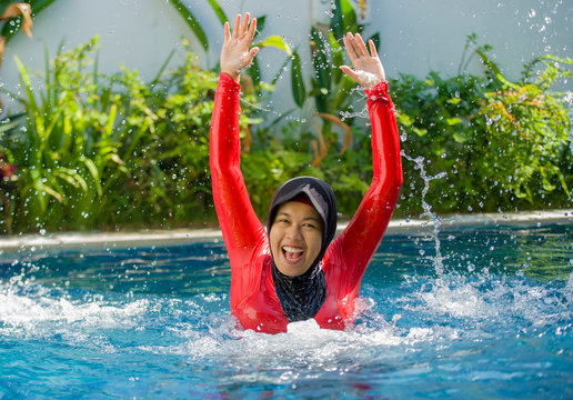 young happy muslim woman playing with water excited in resort swimming pool splashing and having fun wearing traditional islam head scarf islamic swimwear enjoying holidays