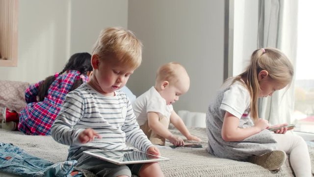 Tracking shot of little children sitting together on bed at home using modern gadgets: boys playing on tablets and girl surfing the Internet on smartphone