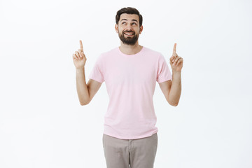 Portrait of charming carefree and delighted male model with dark beard smiling happily looking and pointing up enjoying good day posing in pink t-shirt and pants against gray background