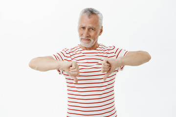 Gloomy displeased mature man with grey beard and hair in striped t-shirt showing thumbs down and...