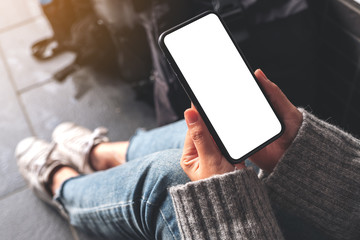 Top view mockup image of a woman holding black mobile phone with blank desktop screen while sitting on the floor with baggage
