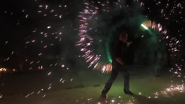Three fire show performers standing in fire circle, performing. One juggles burning sticks, other flails them - slow motion