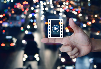 Play button with movie icon on finger over blur colorful night light city with cars in city, Business cinema online concept