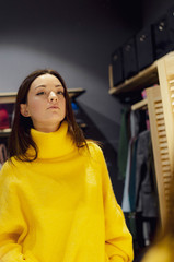 Portrait of attractive caucasian young brunette standing in optician store looking in mirror while trying trendy bright yellow clothes (warm golf or sweater) thinking if she wants to buy them