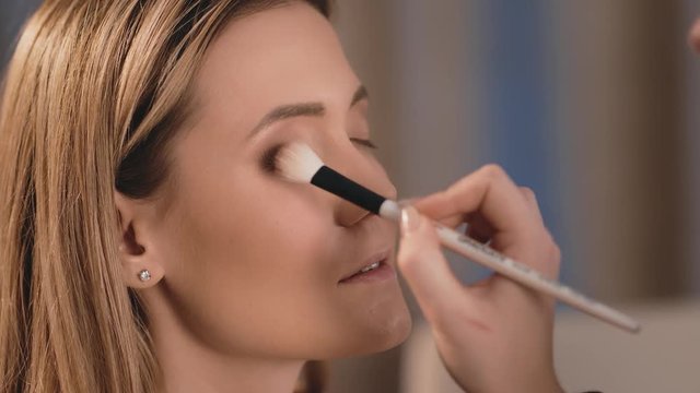 Profile, make-up artist puts dark shadows on the eyelids, professional white brush on the face of a Caucasian blonde model. Close-up, make-up business woman. Slow Motion.