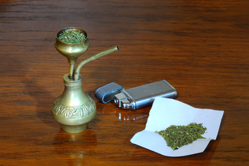 An old metal lighter, a brass bong and a pile of marijuana on a piece of paper – on wooden surface