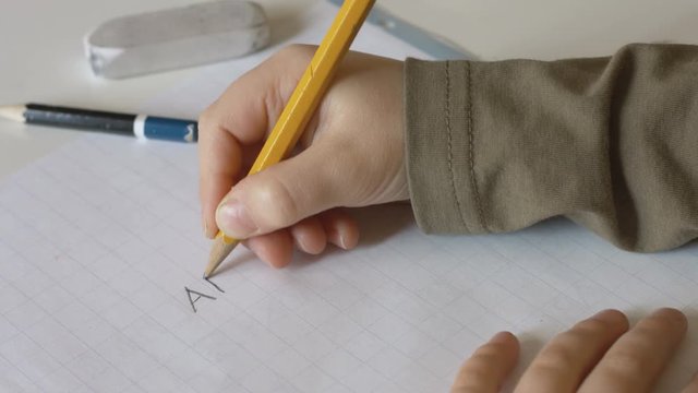 Child writing alphabet letters ABCDE with a pencil on a squared notebook close up