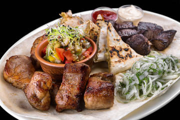 Assorted grilled meat, kebabs, pork, beef, chicken. With pita bread, onions and grilled vegetables. On a white plate. On a black background.