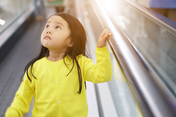 Asian child cute or kid girl down the long escalator alone in department store or supermarket for shopping and looking at the top with wearing yellow shirt on sunlight