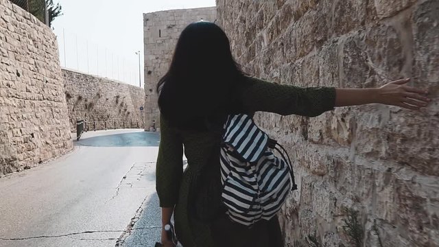 Young brunette with long hair walks down the street of Jerusalem and spending her hand over the stone wall of the old city. Girl tourist in a green dress walks around the city with a selfie stick.