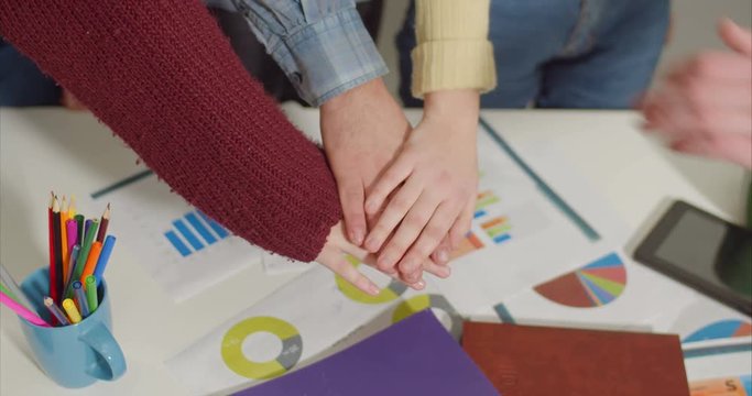Top view of diverse human hands in casual clothes making pile of hands over financial graphs and charts, expressing determination to achieve goals, unity, self motivation to work as successful team.