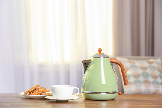 Stylish electrical kettle, cup and plate with cookies on table against blurred room interior. Space for text