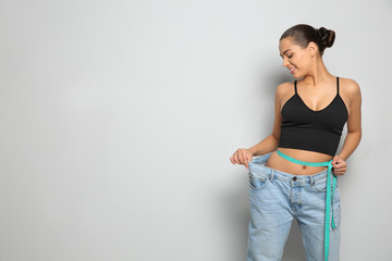 Slim woman in oversized jeans with measuring tape on light background, space for text. Weight loss