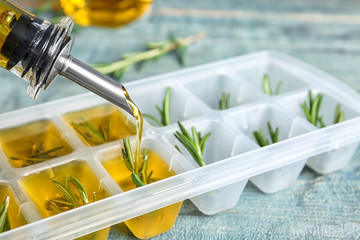 Pouring olive oil into ice cube tray with rosemary on table, closeup