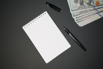 Blank paper spiral notepad with a pen on top on a black background. Dollar banknotes in the corner. 
