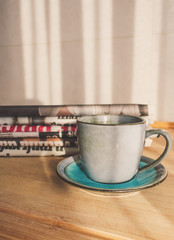 View of a blue cup of coffee on a wooden table with a stack of newspapers to read in the background illuminated by morning sun from a window. Good morning coffee, breakfast and newspapers concept