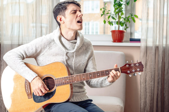 Young attractive male musician sitting on a chair playing acoustic guitar in room. Concept of music as a hobby