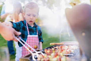 Cute little boy train to turn meat on barbeque grill.Other members of family standing around him...
