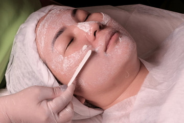 Cosmetic or medical rejuvenating procedure. A beautician or doctor removes the remains of a plaster mask or cream from an Asian woman face in a beauty salon. Close-up.