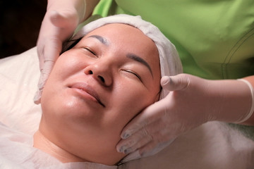 Fototapeta na wymiar A muslim girl with closed eyes is smiling during a face massage at the cosmetology center. Cosmetic rejuvenating facial procedure. Closeup of Asian woman's face and beautician hands in white gloves.