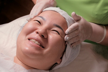 Obraz na płótnie Canvas Cosmetic or medical rejuvenating facial procedure. Close-up face of Asian woman in a beauty salon. Muslim girl smiling during facial massage at the cosmetology center.