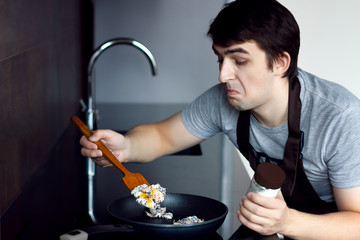 Young loser man male cooking scramble adding salt on metal frying pan in modern loft kitchen. Trying to make breakfast, lunch or dinner. Alone bachelor in kitchen without woman female concept