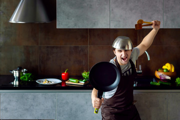 Funny loser man male with metal pan on head frying pan and wooden kitchen utensils in pose of ninja trying to cook failed and yell on grey modern loft kitchen. Unsuccessful bachelor on kitchen concept © Monstar Studio