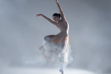 Elegant woman female girl ballerina dancer in beige dress dancing, making performance and dance element ballet in fog dust smoke fume on isolated grey background scene. Dancing in cloud concept