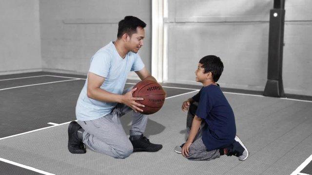 JAKARTA, Indonesia - January 08, 2019: Cute little boy and his father talking together while holding a basketball and sitting in basketball court. Shot in 4k resolution