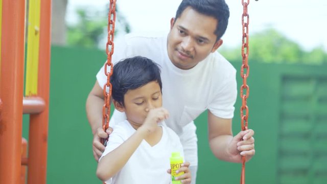 Cute little boy playing soap bubbles with his father while sitting on a swing at playground. Shot in 4k resolution