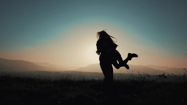 A loving couple runs to meet each other in embrace, man twists his woman against the sunset on the mountains. Slow motion, silhouette