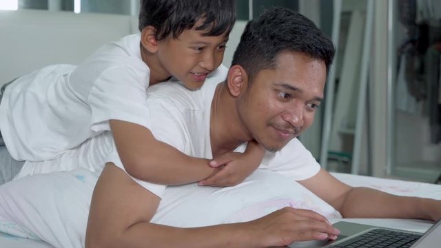 Young father and his cute little boy using a laptop computer while lying on the bed in bedroom at home. Shot in 4k resolution
