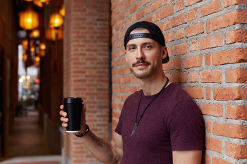 Happy attractive young hip hop man smiling cheerfully while having good conversation, looking at camera, full of energy, holding take away disposable cup with double cappuccino, posing outdoors.