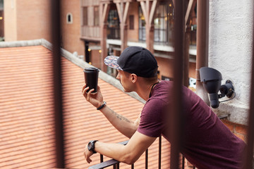 Portrait through stair rails of attractive young Caucasian man in stylish wear enjoying fresh takeaway coffee in paper cup, standing in urban setting over street background. People, lifestyle concept.